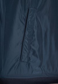 Granatowa puchowa kurtka z poziomą kieszenią na zamek po lewej stronie, o gładkiej, błyszczącej fakturze i prążkowanym wykończeniu.