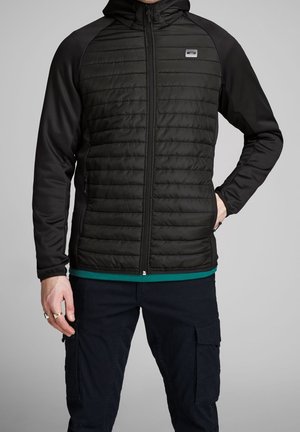 Mann trägt eine schwarze gesteppte Reißverschlussjacke mit Kapuze und schwarze Cargohose, steht mit einer Hand in der Tasche und einer Hand herunter.