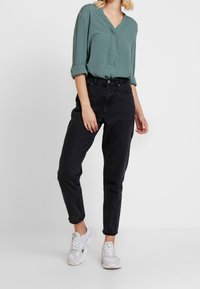 Frau trägt eine gedämpfte grüne V-Ausschnitt-Bluse, schwarze hoch taillierte, schmal zulaufende Jeans und weiße Turnschuhe, steht vor einem weißen Hintergrund.