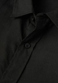 Zwarte shirt met een gestructureerde kraag, voorzien van een knopen sluiting aan de voorkant en een zachte, gestructureerde stof. Close-up zicht benadrukt de details.