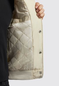 Beige quiltet jakke indvendig med en glat, skinnende tekstur og trykknapper. Nederste kant har en ribbet strikket detalje for ekstra flair.