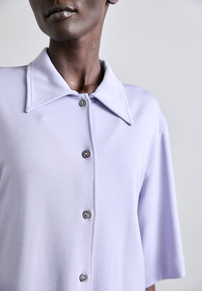 Light lavender shirt with a pointed collar and short sleeves. Five gray buttons on a front placket, smooth texture, minimalistic design.