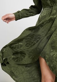 Robe longue vert foncé fluide avec motifs de feuilles tropicales brillantes, manches longues avec poignets, et une fente haute dévoilant une jambe nue.