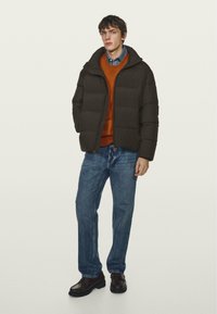Giacca a vento nera con cappuccio, design imbottito, indossata sopra un maglione arancione, abbinata a jeans blu e scarpe scure. Stile semplice e funzionale.