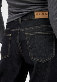 Jeans de mezclilla negra con una parche de cuero marrón etiquetado "NA-KD," que exhibe costuras amarillas y cuatro bolsillos en la parte posterior.