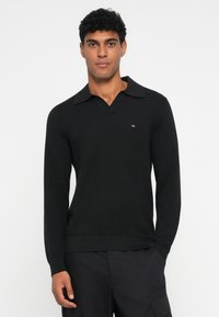 Sort langærmet trøje med v-hals og en struktureret mønster. Har en foldet krave og et lille emblem på brystet.