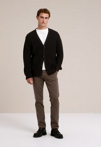 Cardigan negru cu dungi, prevăzut cu cinci nasturi, purtat peste o cămașă albă. Pantaloni maro și pantofi negri completează ținuta. Fundal neutru.