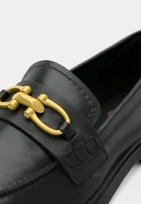 Mocassin en cuir noir avec un accent en métal doré sur le dessus, une couture contrastante et une texture lisse et polie.