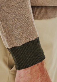 Rękaw swetra z dzianiny, z jasnobrązową górną częścią przechodzącą w ciemnozielony ściągacz, charakteryzujący się gładką teksturą i kontrastującymi kolorami.