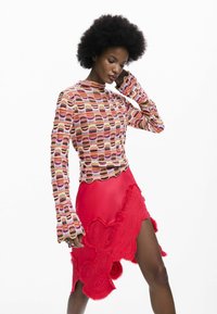 Bluzka z długim rękawem w kolorowy geometryczny wzór; czerwona asymetryczna spódnica z falistymi krawędziami i detalami z fakturowanej haftowanej tkaniny.