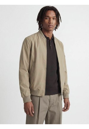 Jeune homme avec des dreadlocks portant une veste bomber beige sur une chemise noire et un pantalon beige, debout devant un fond blanc uni.
