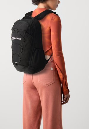 Black backpack with logo, adjustable straps, and elastic cord detailing, worn over an orange long-sleeve top and pink pants.
