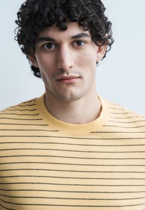 Νέος άνδρας με μαύρα σγουρά μαλλιά που φοράει κίτρινη και μαύρη ριγέ μπλούζα, στραμμένος μπροστά με ουδέτερη έκφραση σε ανοιχτό φόντο.