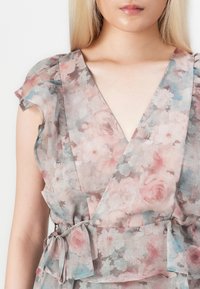 Donna che indossa un vestito avvolgente floreale pastello con maniche corte a volantino e una cintura annodata in vita, che mostra il décolleté e la parte superiore del torso.