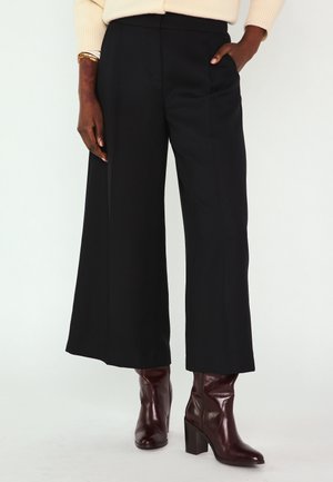 Persona che indossa pantaloni neri a gamba larga e corte, maglione color crema e stivaletti con tacco marrone scuro, in piedi con una mano in tasca.