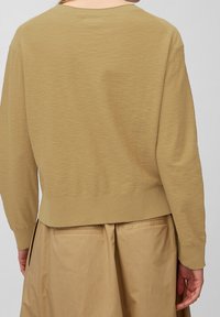 Maglione marrone chiaro lavorato a maglia con scollo rotondo e polsini a coste, caratterizzato da un motivo a trama. Indossato sopra una gonna beige con tasche.