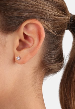 Orecchino a forma di stella argentata con finitura lucida, indossato in un orecchio forato, che mostra linee pulite e un design minimalista.