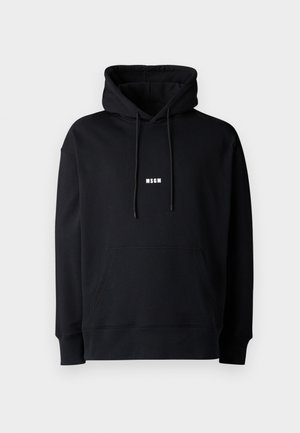 Henkilö pukeutuneena mustaan MSGM-huppariin, jossa etutaskut, mustiin cargohousuihin sivutaskuilla ja mustiin kenkiin, seisoo valkoista taustaa vasten.