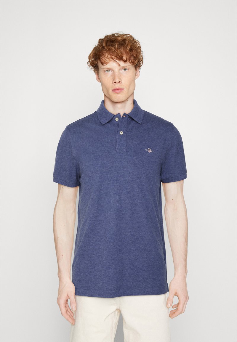 Junger Mann mit lockigem roten Haar, der ein blaues kurzärmeliges Poloshirt und weiße Hose trägt, vor einem schlichten hellen Hintergrund stehend.