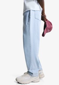 Pantaloni de denim de un albastru deschis, cu o croială lejeră, buzunare laterale și detalii de cusătură subtile, asortați cu adidași albi și o șapcă vișinie.