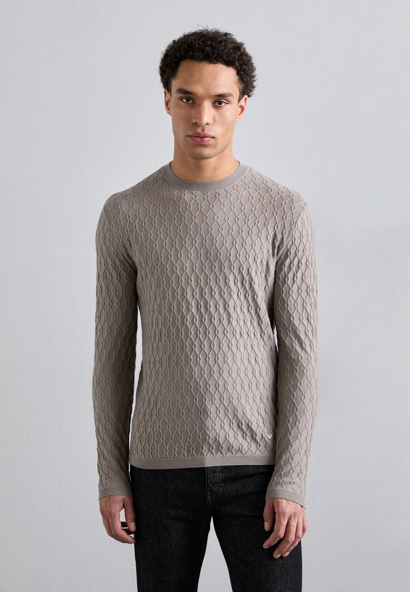 Gai pelēks adīts džemperis ar garām piedurknēm, kas piedāvā teksturētu dimanta rakstu, apaļu dekoltē un ribota apdare pie piedurknēm. Kombinēts ar tumši ziliem džinsiem.