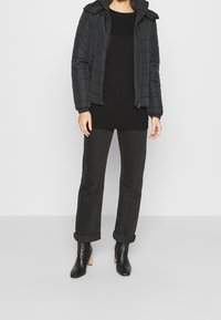 Chaqueta acolchada negra con cierre de cremallera y cuello plegado, llevada sobre una blusa larga negra, combinada con pantalones oscuros remangados y botas negras de tacón.