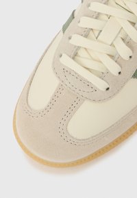 Prim-plan cu vârful unei adidași din piele intoarsă bej și piele crem, cu șireturi crem și talpă texturată din gumă, pe fundal alb.