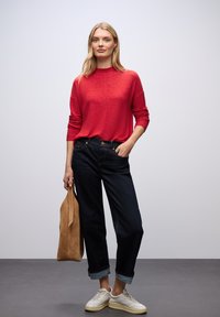 Czerwona sweter z długim rękawem, ciemnoniebieskie jeansy o luźnym kroju, beżowa torebka z zamszu i białe sneakersy z teksturowanymi akcentami. Prosty, codzienny strój.