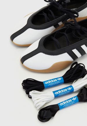 Par hvide og sorte Adidas sko med gummisåler placeret tæt på tre sæt sorte, hvide og sorte satin skosnørebånd med blå Adidas-mærker.