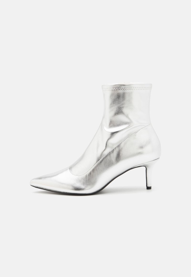 Bota de tobillo metálica plateada con punta afilada y tacón delgado. Presenta un ajuste ceñido y una textura suave y brillante. Detalles de costura en la parte superior.