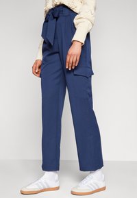 Navy blue high-waisted trousers with a belted waist, side pockets, and wide-leg design, paired with white sneakers. Soft fabric texture.