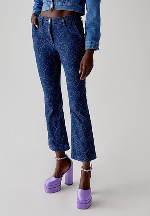 Pantaloni a zampa d'elefante in denim blu scuro con ricami floreali e chiusura con bottone, abbinati a scarpe con tacco a piattaforma viola.