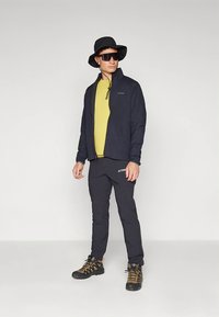 Veste marine avec fermeture éclair et cordon de serrage, chemise jaune, pantalon noir avec logo, casquette noire, et chaussures de randonnée avec accents jaunes.