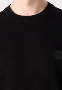 Gros plan d'une personne portant un pull en maille noir à col rond avec un petit écusson logo rectangulaire noir sur la poitrine.