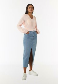 Hellrosa Bluse mit langen Ärmeln und V-Ausschnitt, mit gerafften Ärmeln, kombiniert mit einem mittellangen, hellblauen Jeansrock mit einem Schlitz vorne. Weiße Sneakers.
