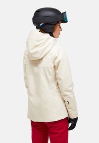 Person iført en cremet skijakke, røde skibukser, sorte handsker og en sort skihjelm med goggles, vendt mod højre på en hvid baggrund.