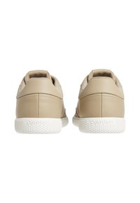 Zapatillas de cuero beige con una textura suave, parte trasera redondeada y suelas de goma blancas. Presentan un sutil logo "Calvin Klein" en el talón.