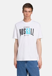 White cotton t-shirt featuring black and teal graphic text "GEORGE RUS63LL." Finished with a crew neck and short sleeves, paired with blue denim jeans.