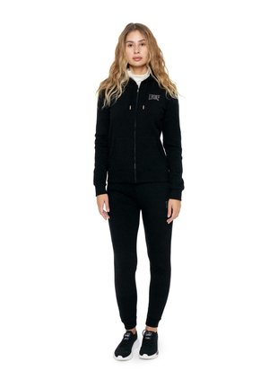 Una mujer luce una sudadera negra con capucha y cremallera, pantalones de chándal a juego y zapatillas negras sobre un fondo blanco.