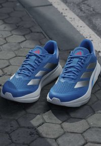 Paire de chaussures de course athlétiques bleues avec semelles blanches et bandes argentées posées sur un pavage hexagonal en pierre près d’un trottoir.