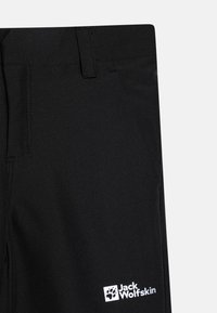 Svarta utomhusbyxor med en sidficka och en vit Jack Wolfskin-logotyp nära fållen, visade mot en enkel bakgrund.