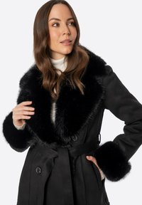 Abrigo negro con un cuello y puños de piel sintética. Presenta una cintura ceñida con cinturón y botones. La tela tiene una textura suave y un diseño ajustado.