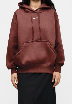 Persoon die een kastanjebruine Nike-hoodie draagt met voorzak en trekkoorden, gecombineerd met een zwarte rok, staand tegen een effen achtergrond.