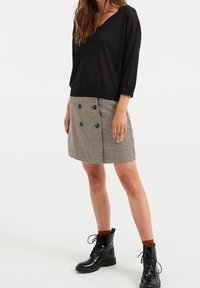 Suéter negro semitransparente de punto con escote en V y mangas de 3/4, combinado con una falda corta de cuadro de perro de paja en beige y negro que presenta botones dobles. Botas de combate negras.