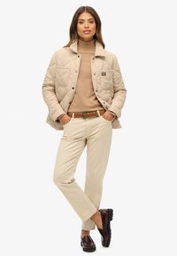 Beige gesteppte Jacke mit aufgenähter Tasche, brauner Rollkragenpullover, beigefarbene Hose, brauer geflochtener Gürtel und bordeauxfarbene Slipper mit strukturierten Sohlen.