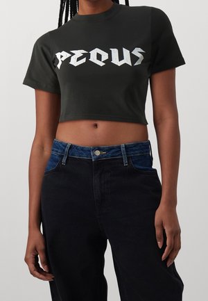 Osoba ubrana w czarny krótki T-shirt z napisem "PEQUIS" oraz wysokim stanem czarne dżinsy z niebieskim detalem w pasie.