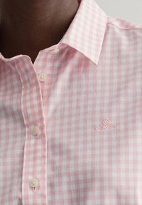 Close-up van een roze gingham overhemd met een knoopsluiting op de kraag en een geborduurd logo op de borst. De stof lijkt lichtgewicht en casual.