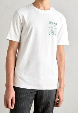 T-shirt en coton blanc avec manches courtes et col rond. Il présente un imprimé graphique vert sur le côté gauche de la poitrine. Texture subtile.
