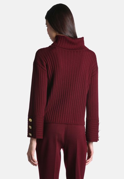 Femme portant un pull bordeaux côtelé à manches longues avec un col montant et des boutons dorés aux poignets, assorti à un pantalon coordonné.