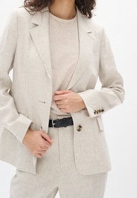 Světle béžový lněný blazer se strukturovanými klopami, s jedním knoflíkem na zapínání a černým pásem. Texturovaná látka s uvolněným střihem.